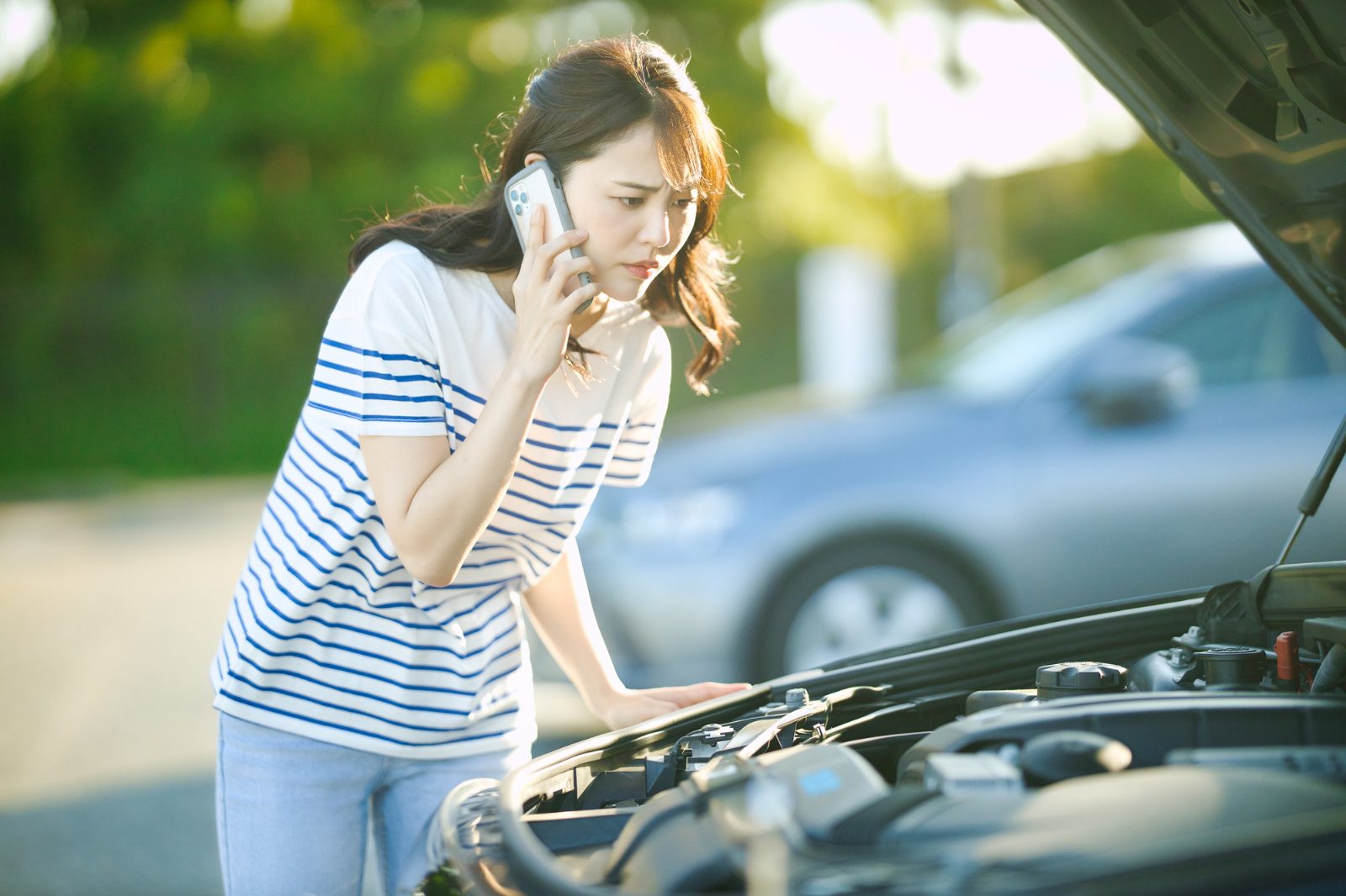 ボンネットを開けた車の前でスマートフォンで連絡する女性。社用車の故障で急に代車が必要になった場面